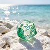 A green polyhedral dice set resting on white rocks near clear blue ocean water.