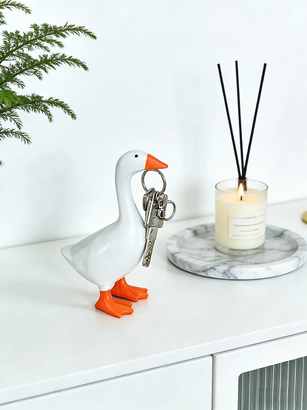 A white goose sculpture with an orange beak and legs stands next to a glass candle holder and a set of black sticks on a marb