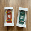 A wooden fingerboard skateboard with realistic trucks is displayed in a white box on a wooden surface.