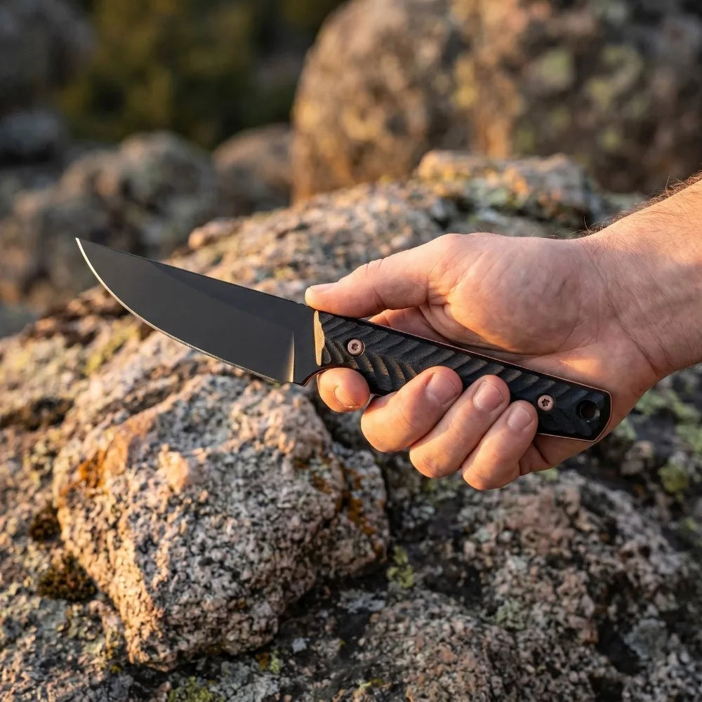 A hand holds a full tang fixed blade knife with a black blade and G10 handle against a rocky background.