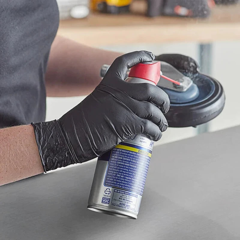 A hand wearing a black nitrile glove is holding a spray bottle of cleaning solution.