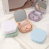 A pink compact contact lens travel case is shown open with a mirror and storage compartments, alongside a white and light gre