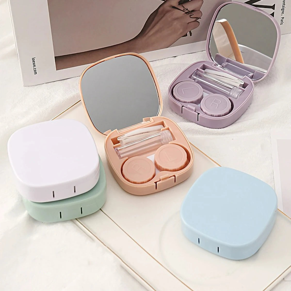 A pink compact contact lens travel case is shown open with a mirror and storage compartments, alongside a white and light gre
