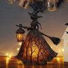 A dark metal witch lantern shaped like a woman with a broom, glowing warmly on a wooden surface.