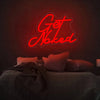 A glowing red neon sign spelling out Get Naked mounted on a dark wall above a bed.