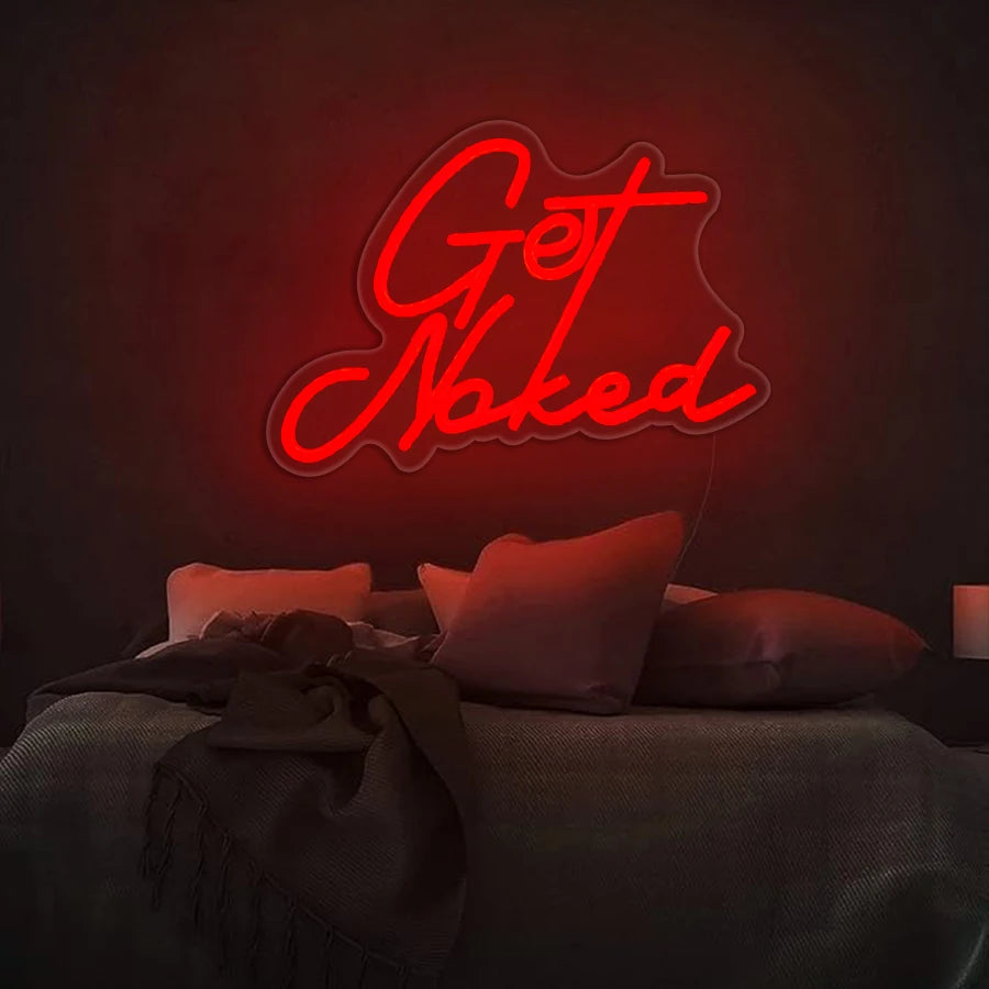 A glowing red neon sign spelling out Get Naked mounted on a dark wall above a bed.