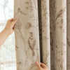 A close-up of a beige blackout curtain panel featuring a brown plant pattern, held by a hand.