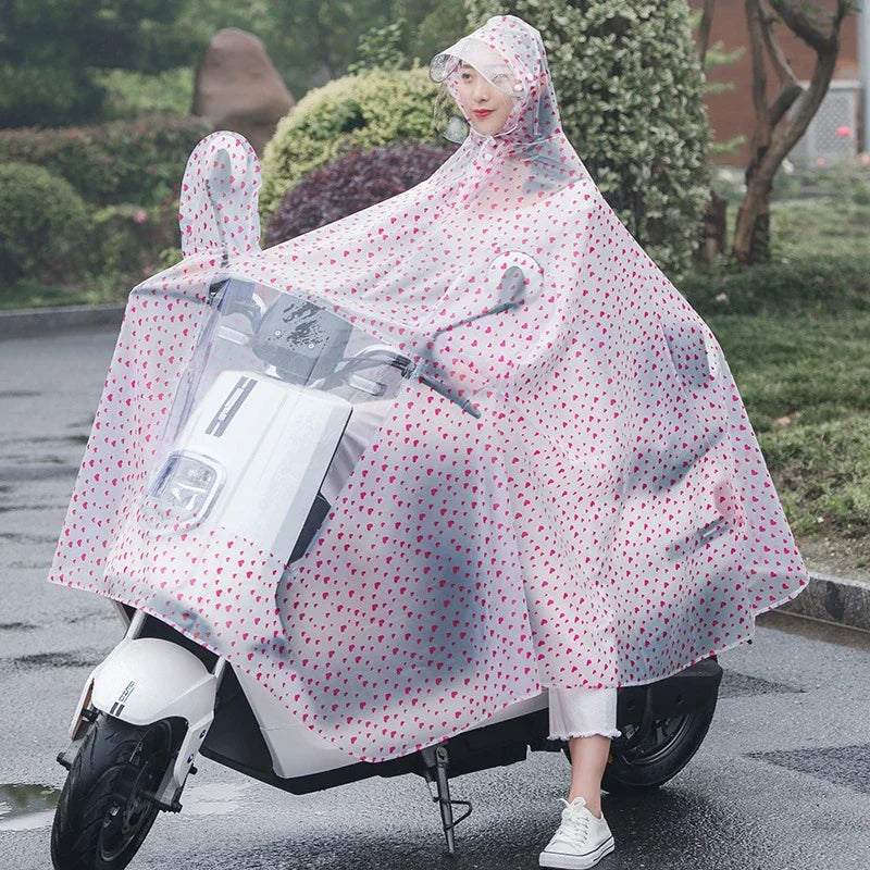A woman wearing a transparent pink polka-dot waterproof motorcycle rain poncho with a hood parked on wet asphalt.