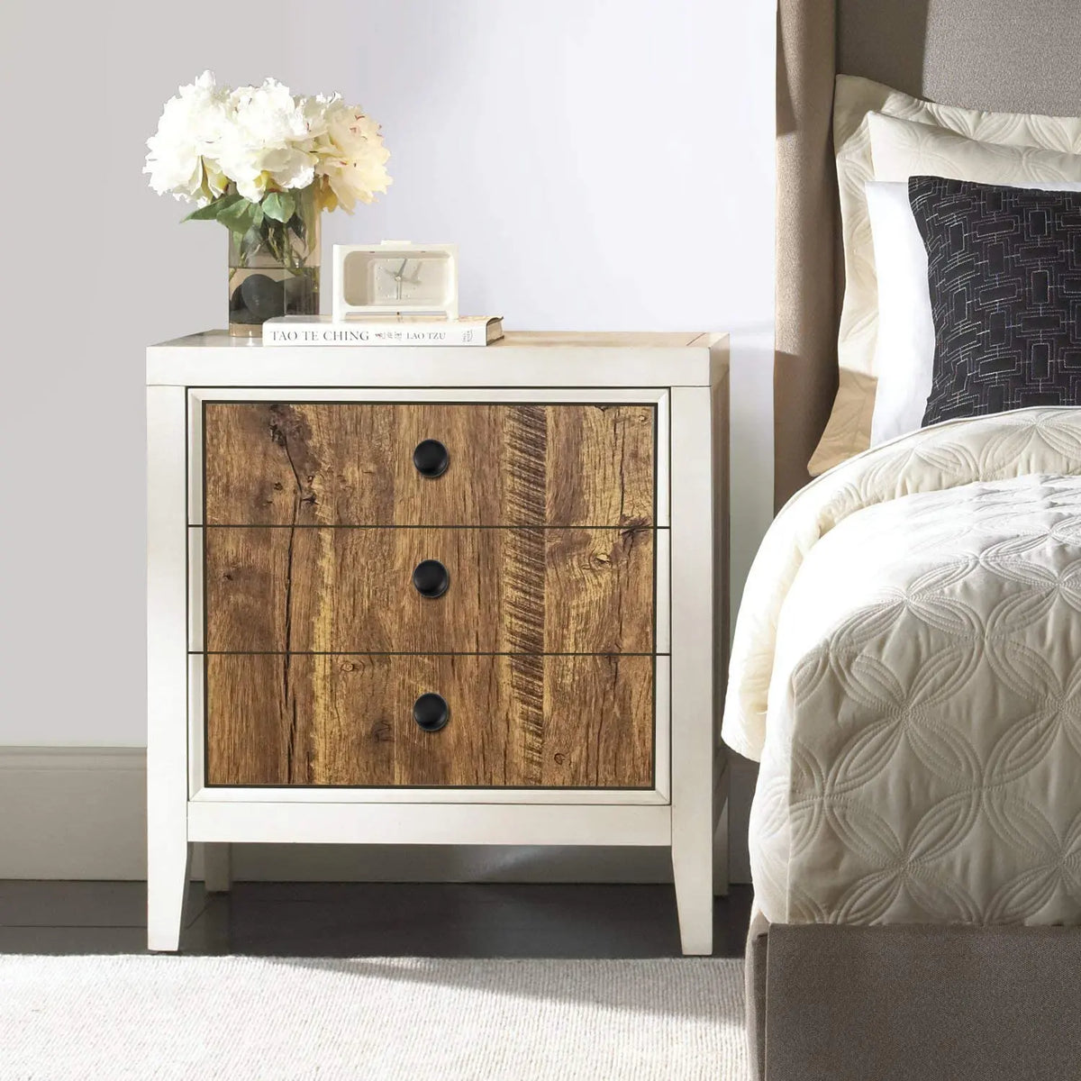 A white wooden dresser with a wood grain drawer front, topped with white flowers and a book.