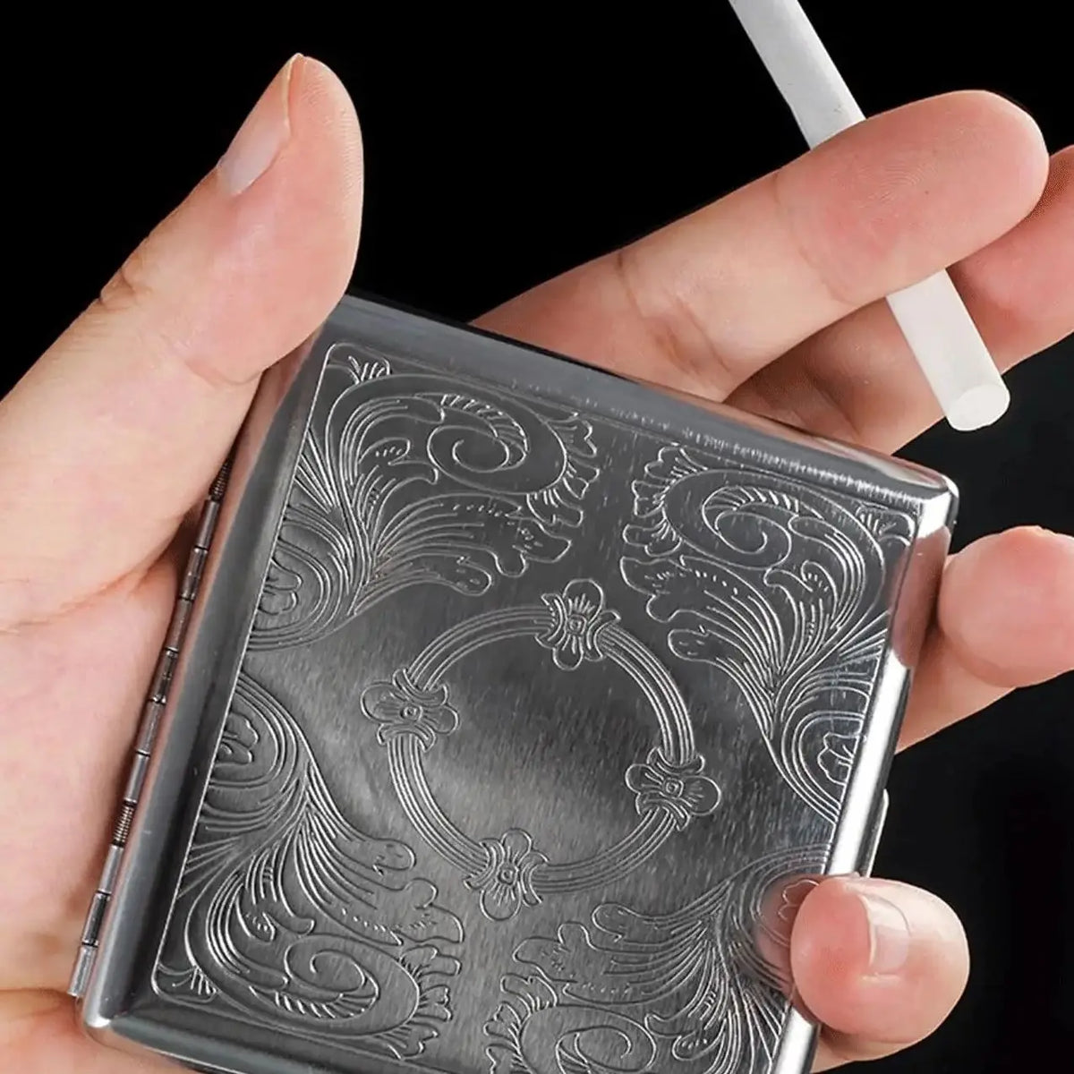 A hand holds a classic stainless steel cigarette case with an embossed floral pattern.