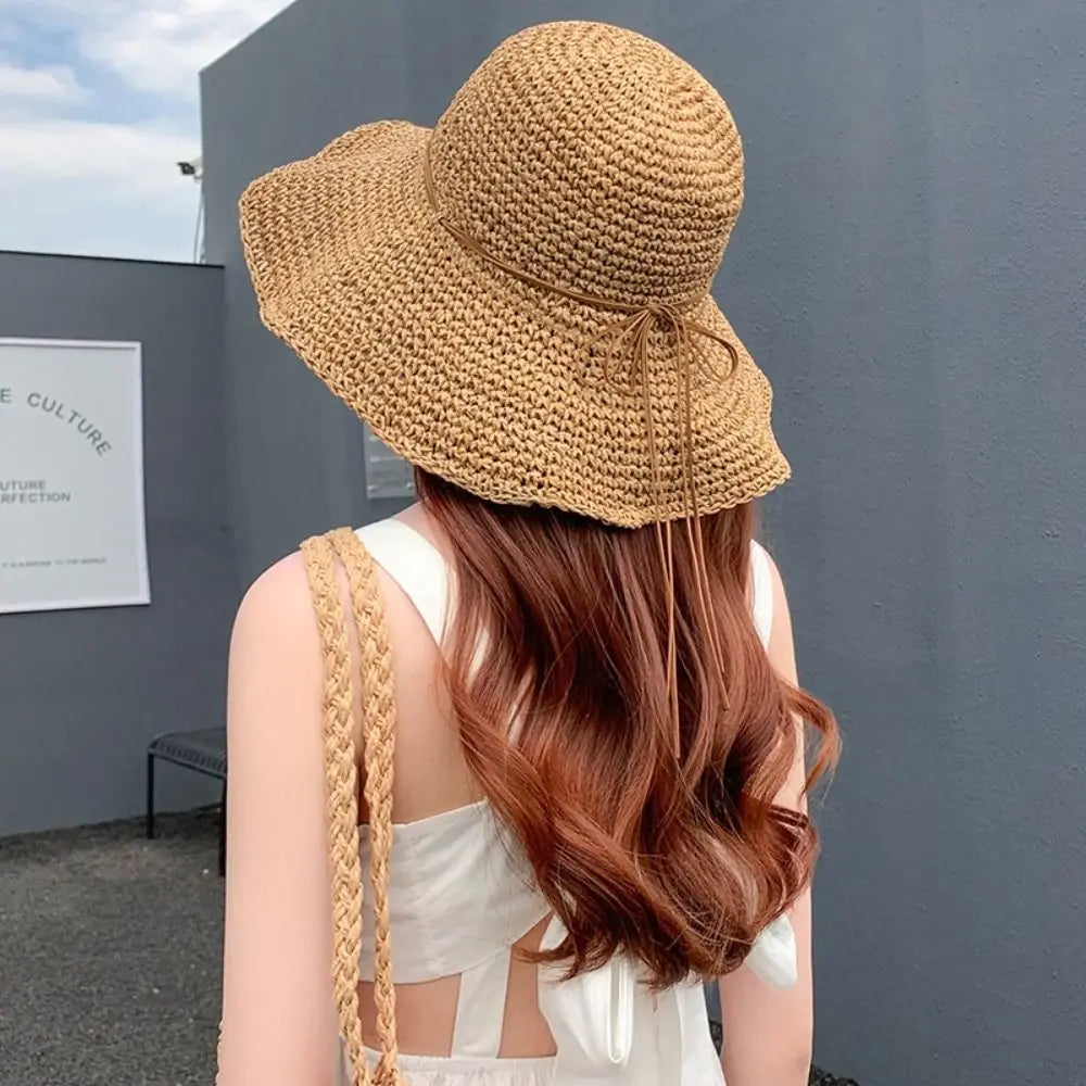 Una mujer con sombrero de paja de ala ancha y una blusa blanca, vista desde atrás contra una pared gris.