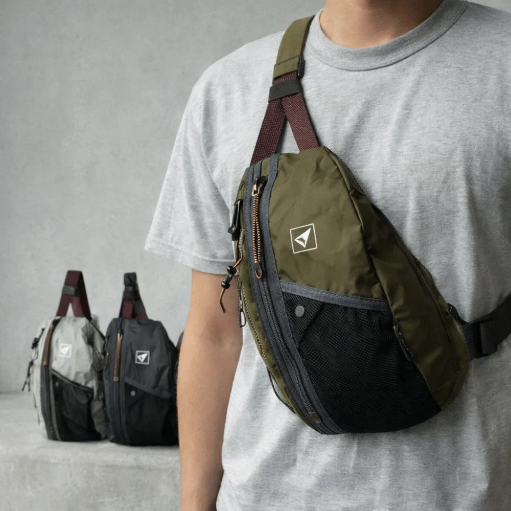 A person wearing a gray t-shirt holds an olive green nylon crossbody bag with a dark strap.