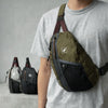 A person wearing a gray t-shirt is holding an olive green and black nylon crossbody bag with a brown adjustable strap.