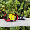 A pair of red and black HD Motocross goggles with a yellow anti-fog lens resting on a gray surface amidst green foliage.