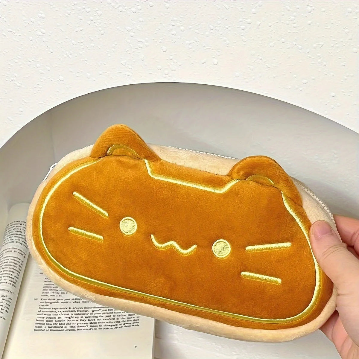 A fluffy orange cat plush pencil case is shown resting on a white surface.
