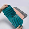 A hand holds a teal silicone faucet mat and sink splash guard alongside two other mats.