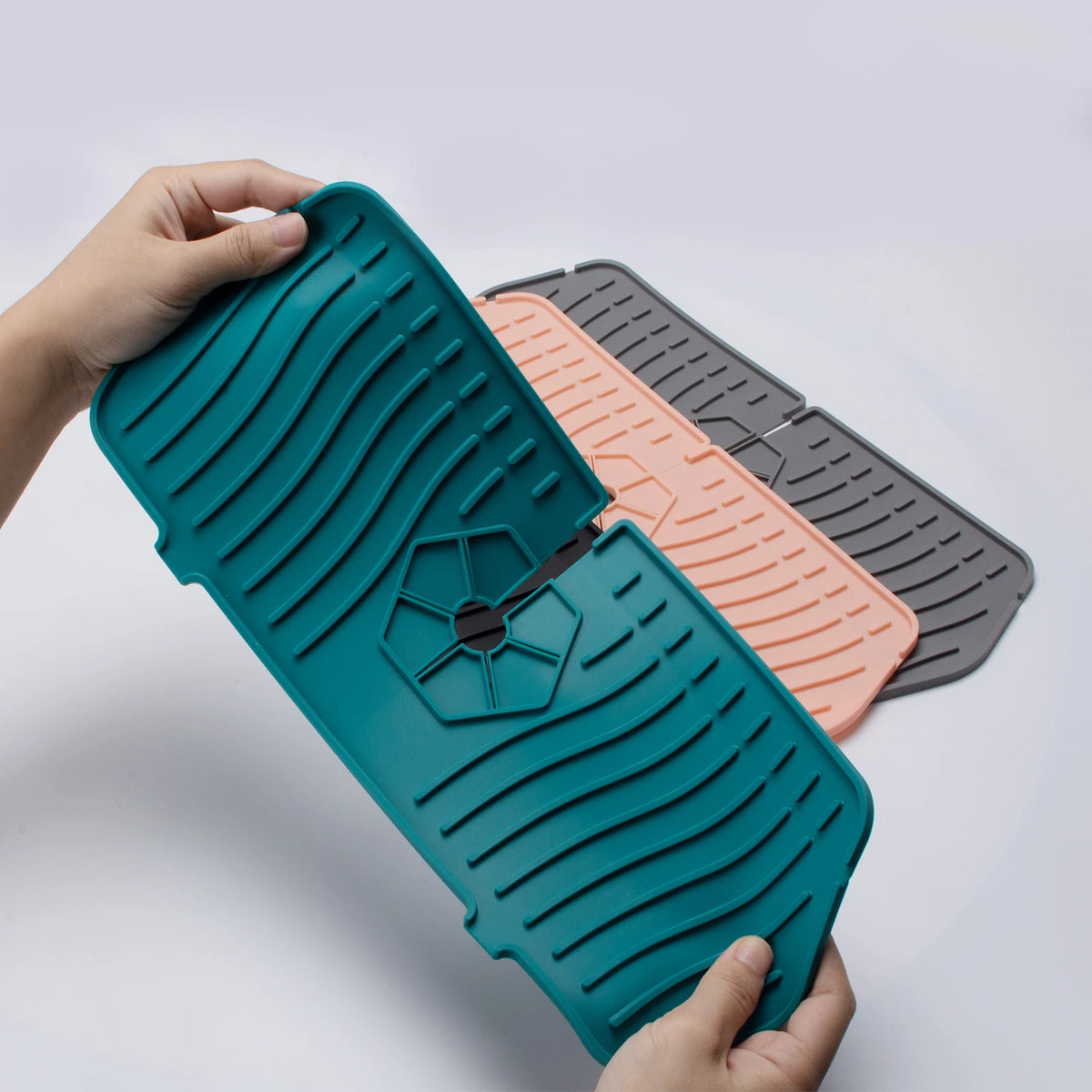 A hand holds a teal silicone faucet mat and sink splash guard alongside two other mats.