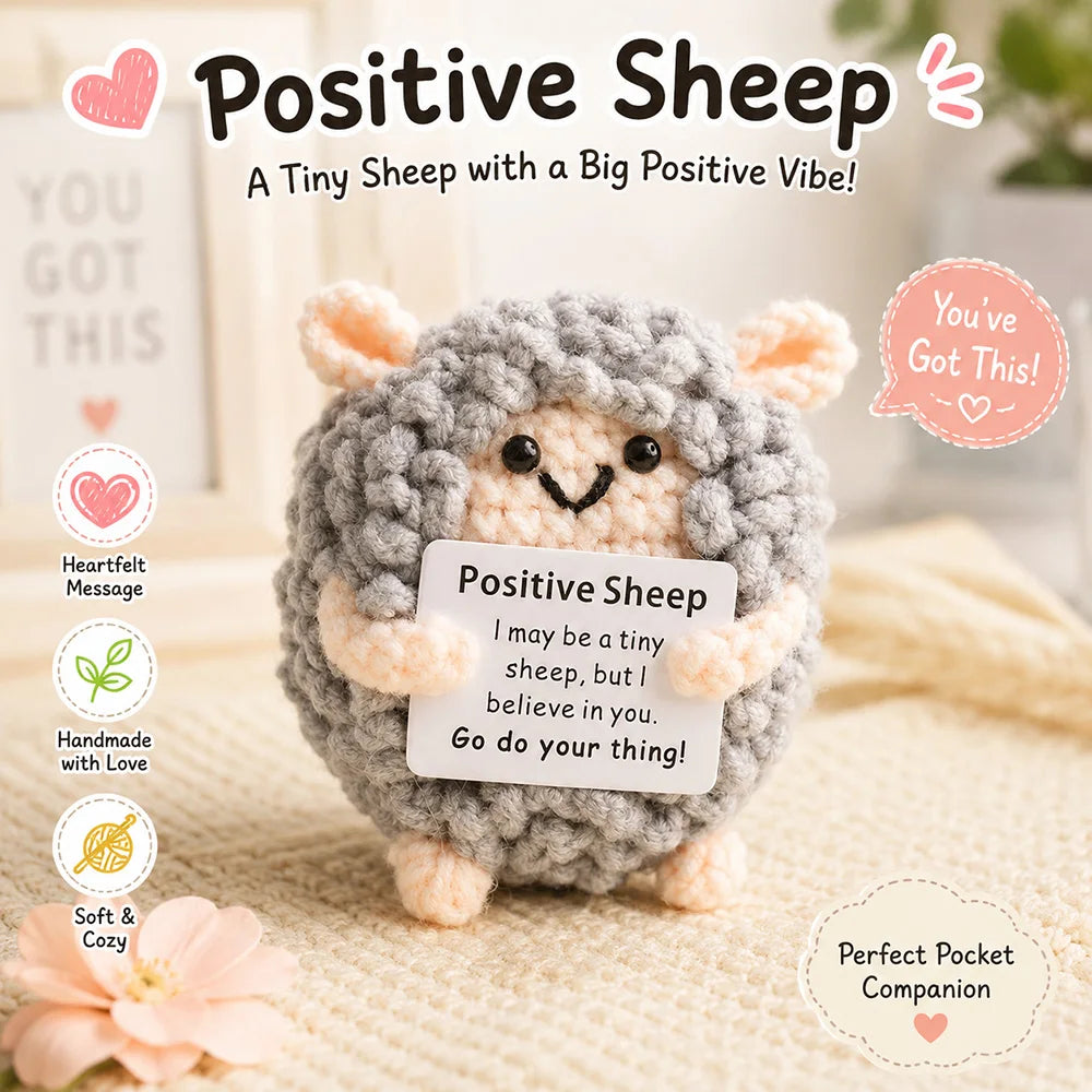 A handmade grey crochet sheep plush toy with a smiling face and a positive message tag.