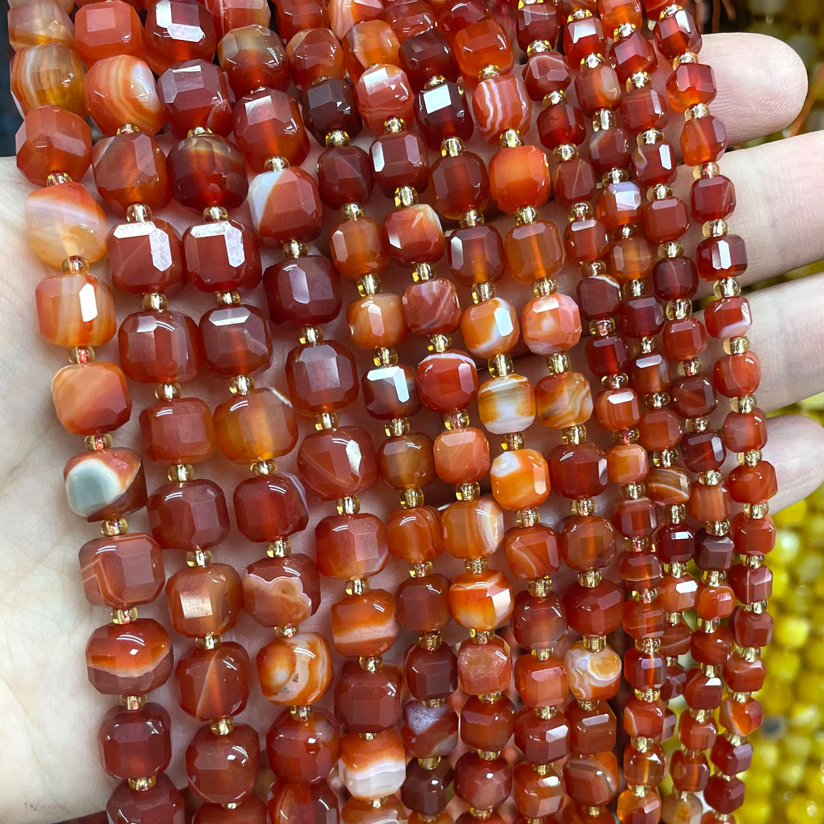 A hand holds a string of square faceted natural stone beads in various shades of reddish-brown and amber for jewelry making