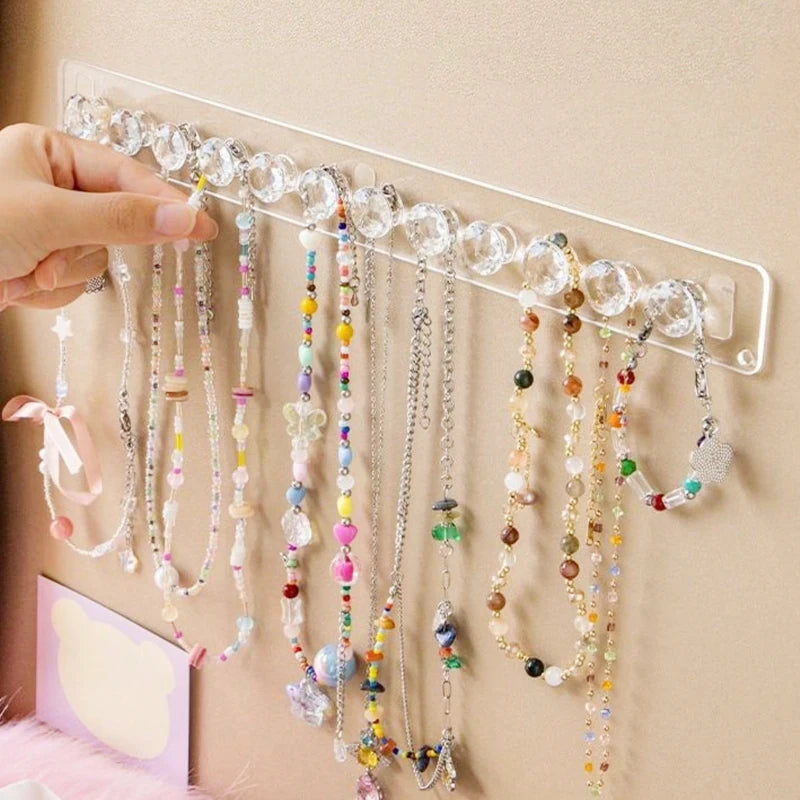A hand is displaying a wall-mounted acrylic jewelry organizer with crystal knobs and various beaded necklaces.
