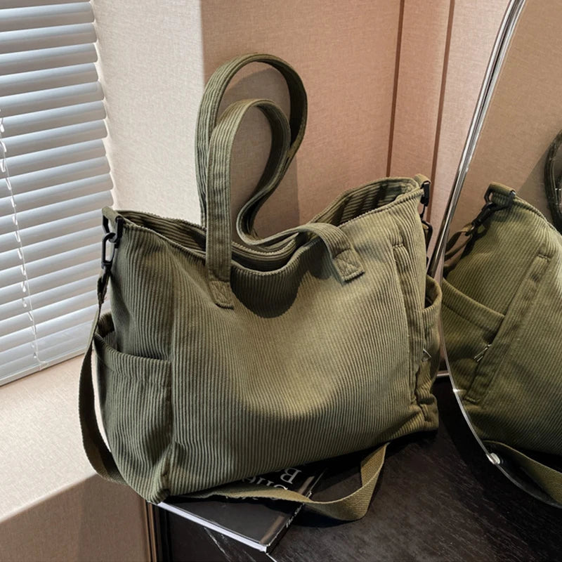 A olive green corduroy tote bag with a fabric strap is displayed next to a window.
