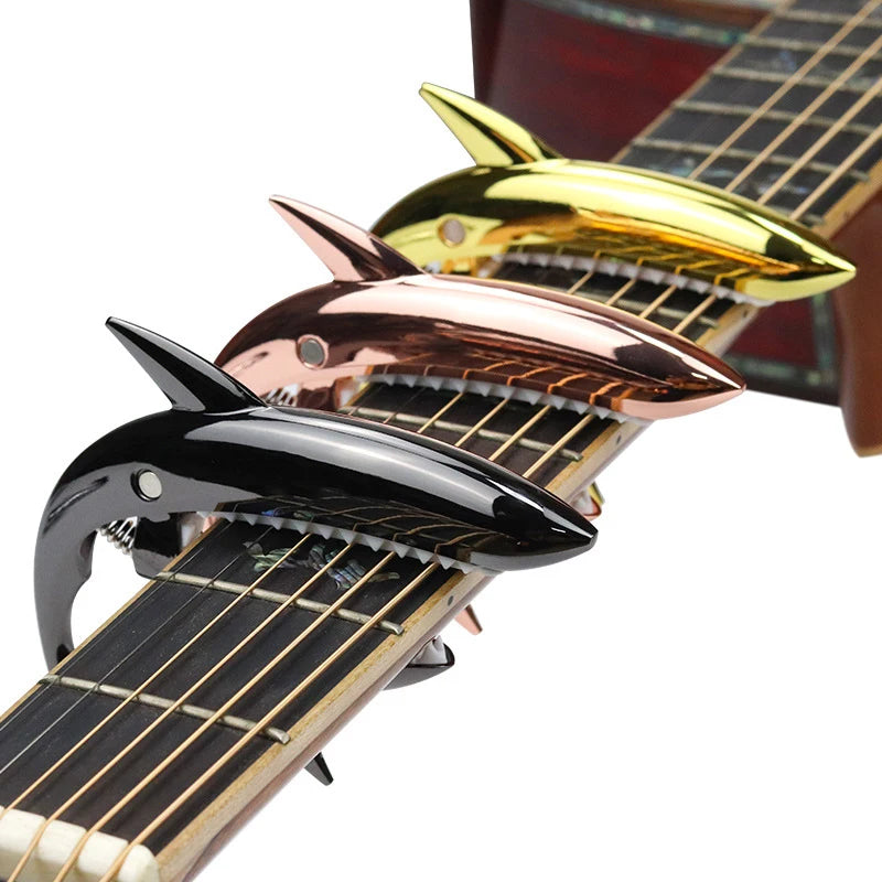 A close-up of a guitar capo featuring three shark-shaped metal pieces in gold, rose gold, and black.
