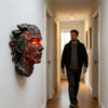 A dark, cracked stone sculpture resembling a human head with glowing orange internal light mounted on a white wall.