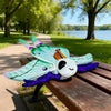 A plush toy of a Luna moth with green and purple wings resting on a wooden bench in a park setting.