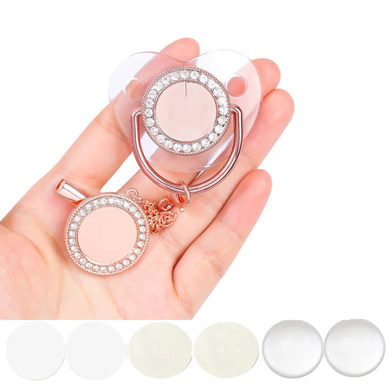 A hand holds a sparkling rhinestone pacifier with a clip for babies, featuring a pink silicone ring and a clear dome.