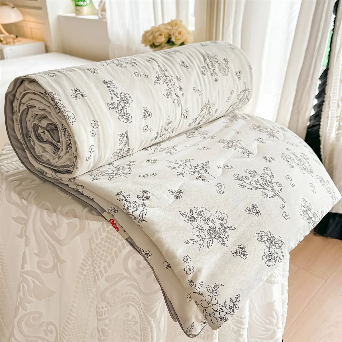 A rolled white quilt with a floral print is displayed on a bed, next to sheer curtains.