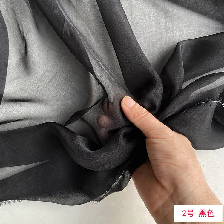A hand is folding a piece of black sheer silk chiffon fabric to show its drape and texture.