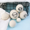 A set of six white wool dryer balls are displayed in a black wire basket on a light blue surface.
