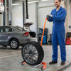 A man in blue overalls is holding an orange tire dolly with a mounted tire in a garage setting.