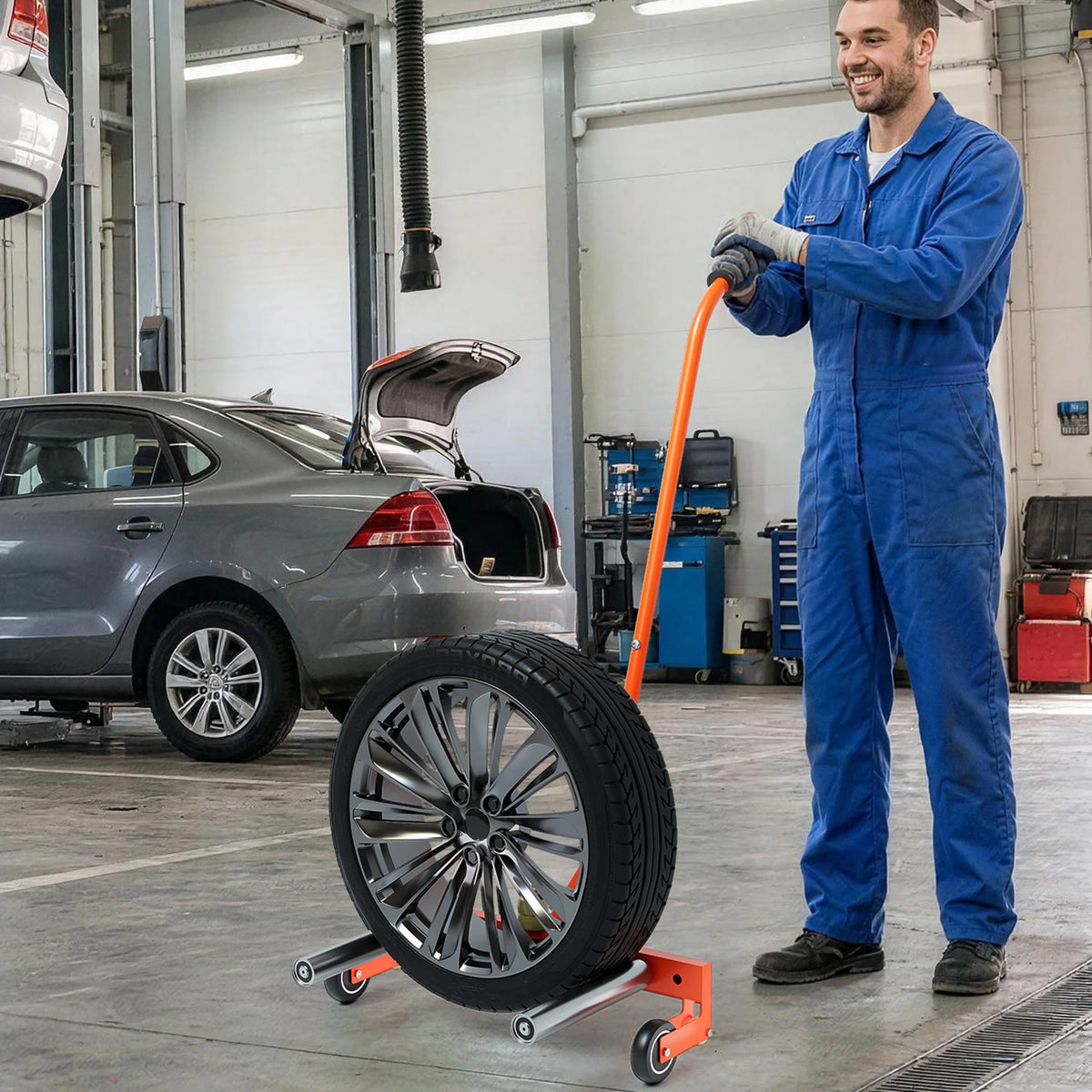 A man in blue overalls is holding an orange tire dolly with a mounted tire in a garage setting.