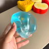 A hand holds a translucent blue squishy stress relief toy next to yellow and red fruit shapes.
