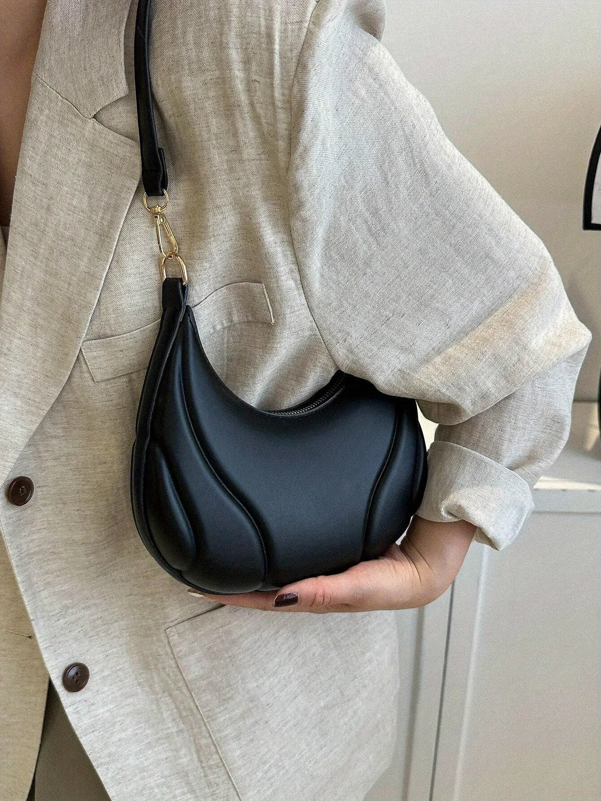 A woman holds a black textured crossbody bag with a gold chain strap against a light beige blazer.