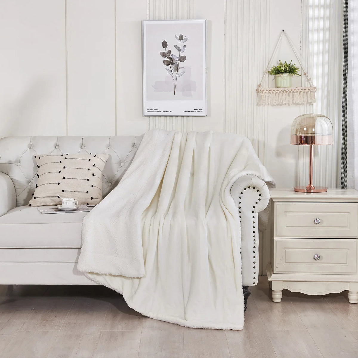 Una manta suave de sherpa crema drapeada sobre un sofá blanco en una sala de estar luminosa y moderna.