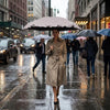 A woman in a beige trench coat holds a large black umbrella while walking on a wet city street.