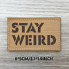 A tan woven patch with black text that reads STAY WEIRD mounted on a wooden surface.
