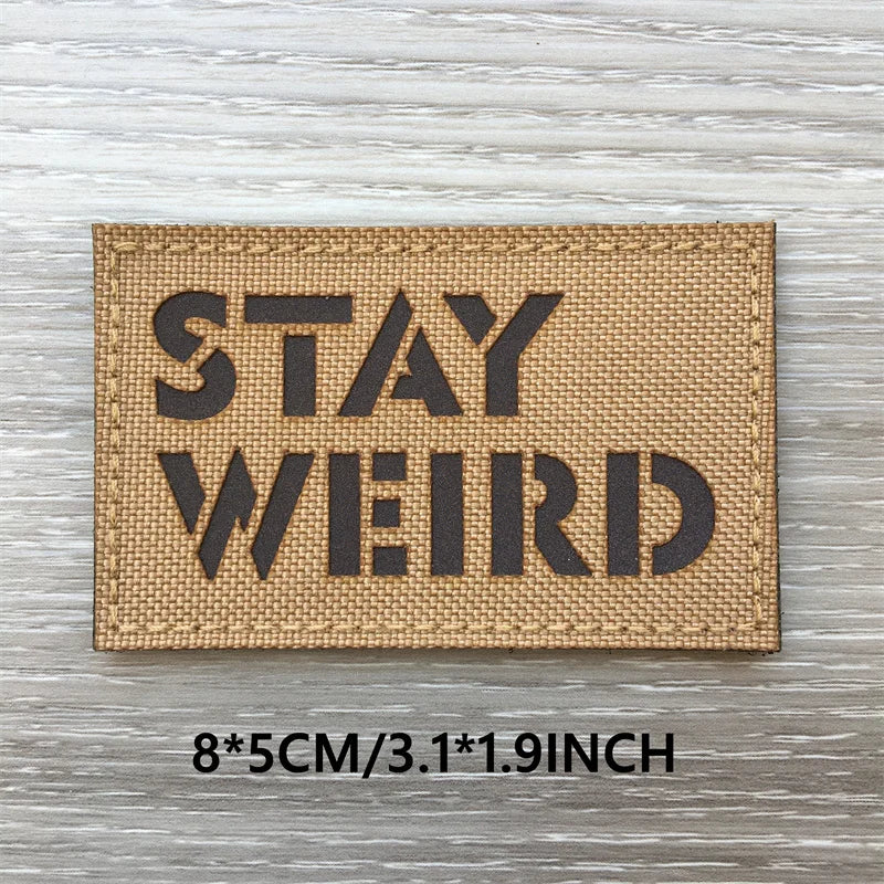 A tan woven patch with black text that reads STAY WEIRD mounted on a wooden surface.