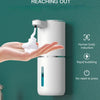 A hand is reaching out to a white touchless soap dispenser, which is producing a bubble immediately upon contact.