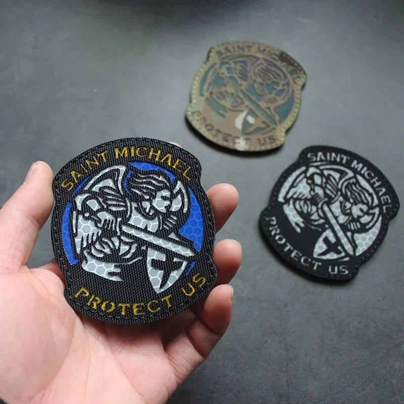 A hand holds a black reflective patch featuring the text Saint Michael Protect Us and an archangel design.
