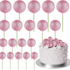 Eine Sammlung von 20 rosa Glitzer-Ball-Kekstoppern auf grünen Stäbchen um einen weißen Kuchen arrangiert.