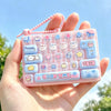 A hand holds a pink acrylic keycap fidget keychain with various cartoon-themed buttons and a pink beaded chain against a brig