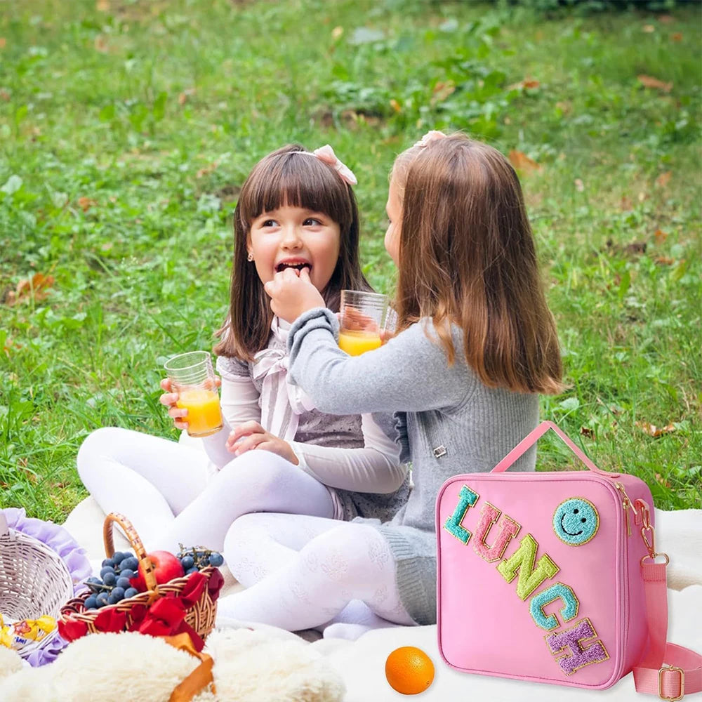 A young girl is holding an orange juice glass while another girl looks at her, sitting outdoors on a blanket with a pink insu