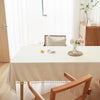 A light beige rectangle dining table covered with a linen tablecloth and cotton tassels in a bright room.