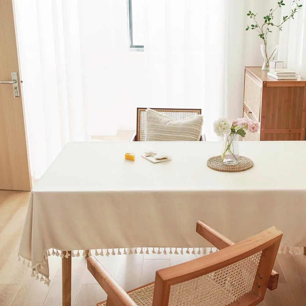 A light beige rectangle dining table covered with a linen tablecloth and cotton tassels in a bright room.