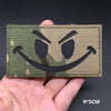 A close-up of a green camouflage tactical patch featuring a black menacing face design being held in a hand.
