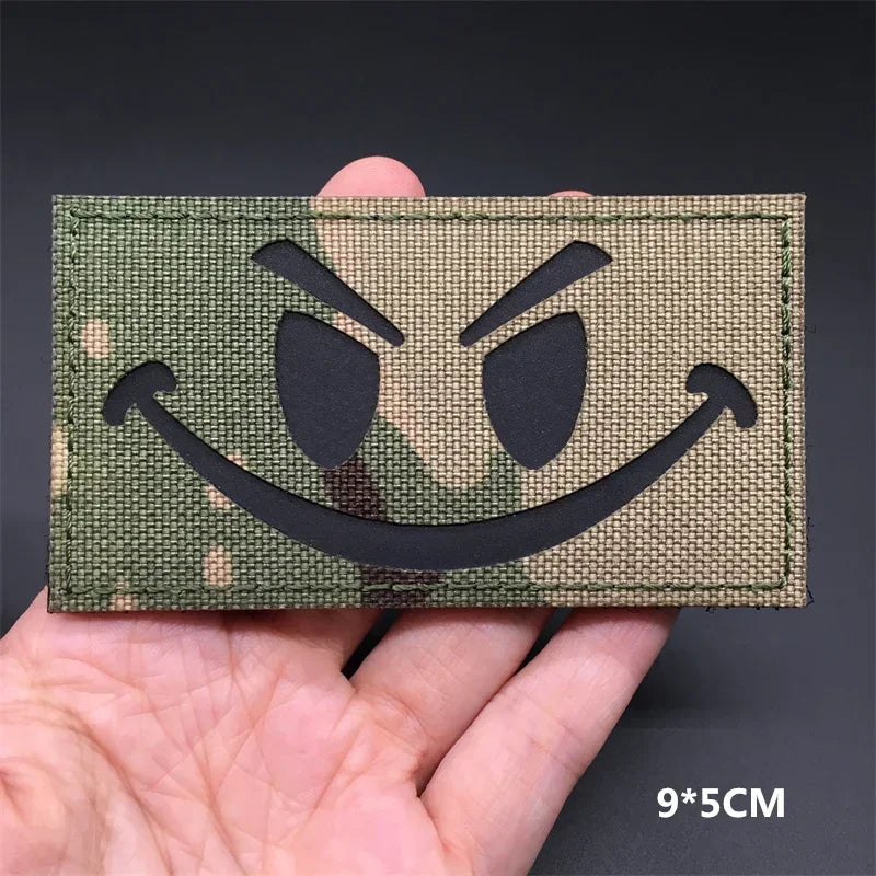 A close-up of a green camouflage tactical patch featuring a black menacing face design being held in a hand.