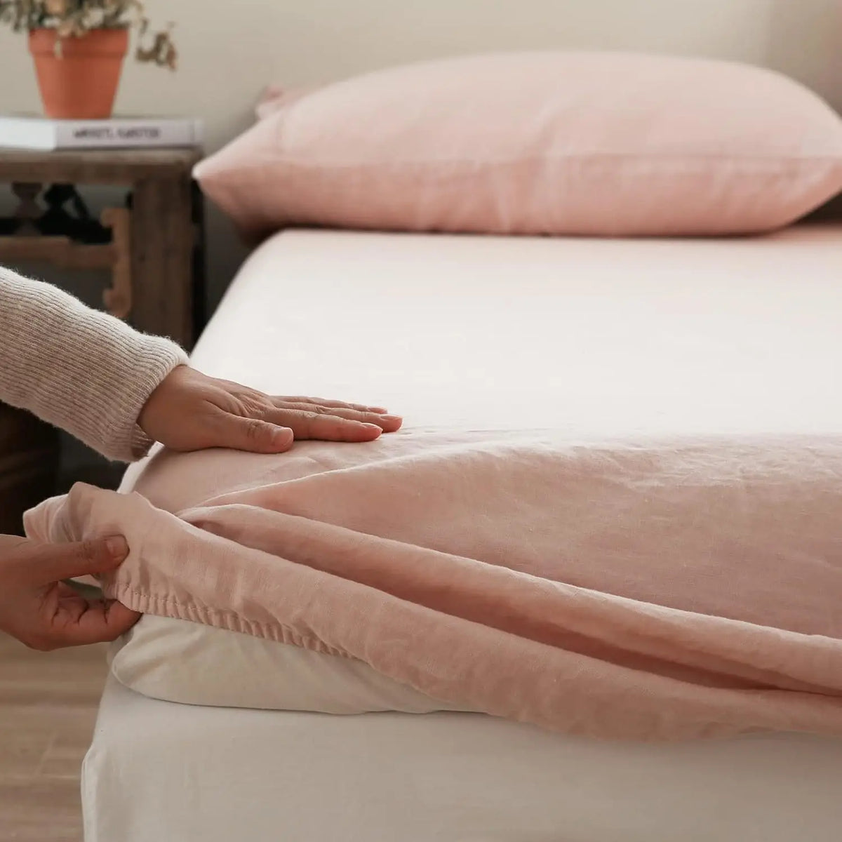 A close-up of a pink linen fitted sheet on a bed with a hand adjusting the fabric.